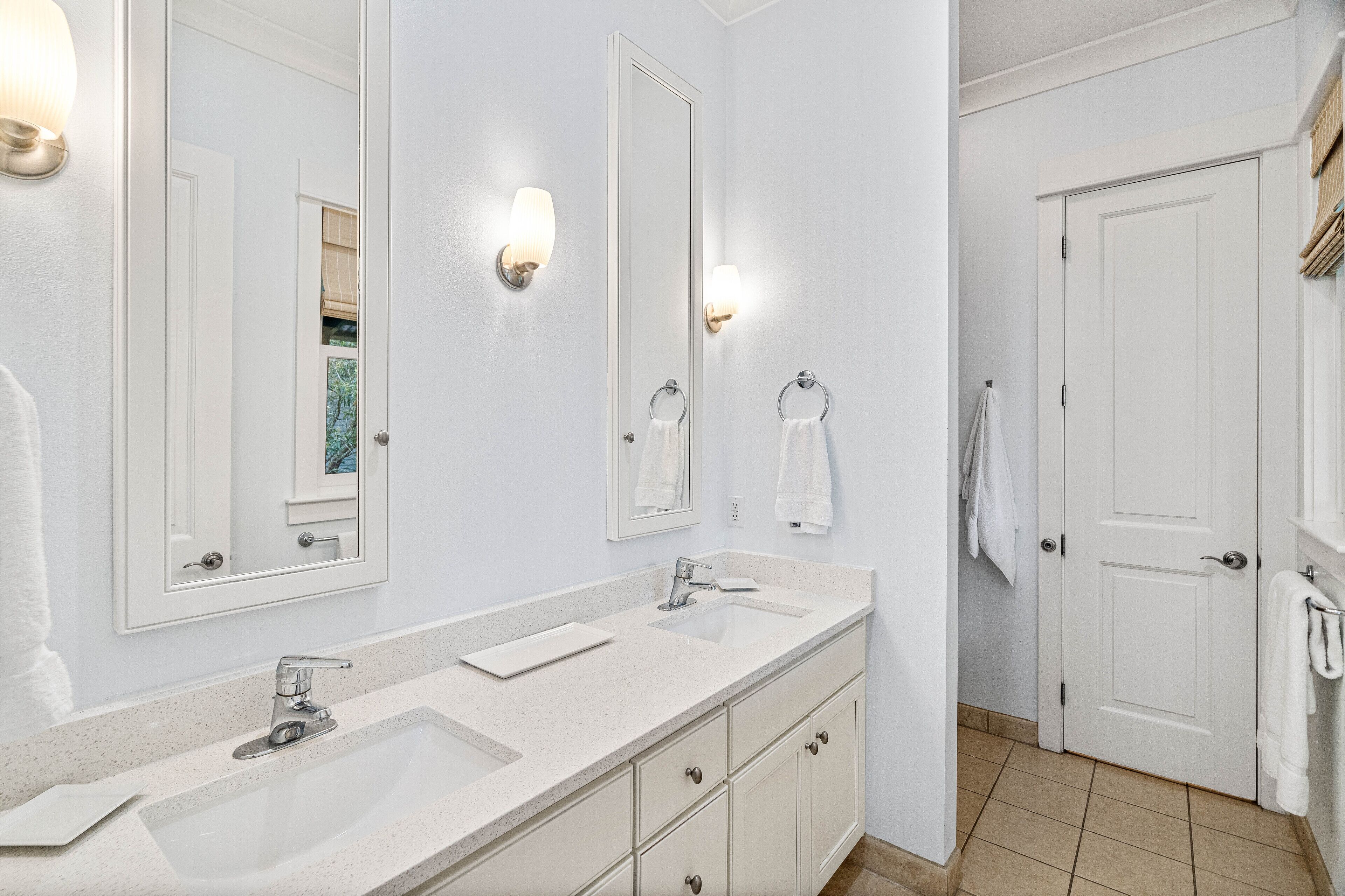 A bathroom with two sinks, a large mirror, wall sconces, and a door with a towel rack.