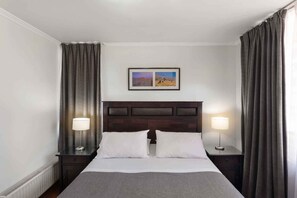 Quarto de hotel com uma cama grande, duas mesas de cabeceira com abajures, uma cabeceira escura e uma moldura com um quadro na parede.