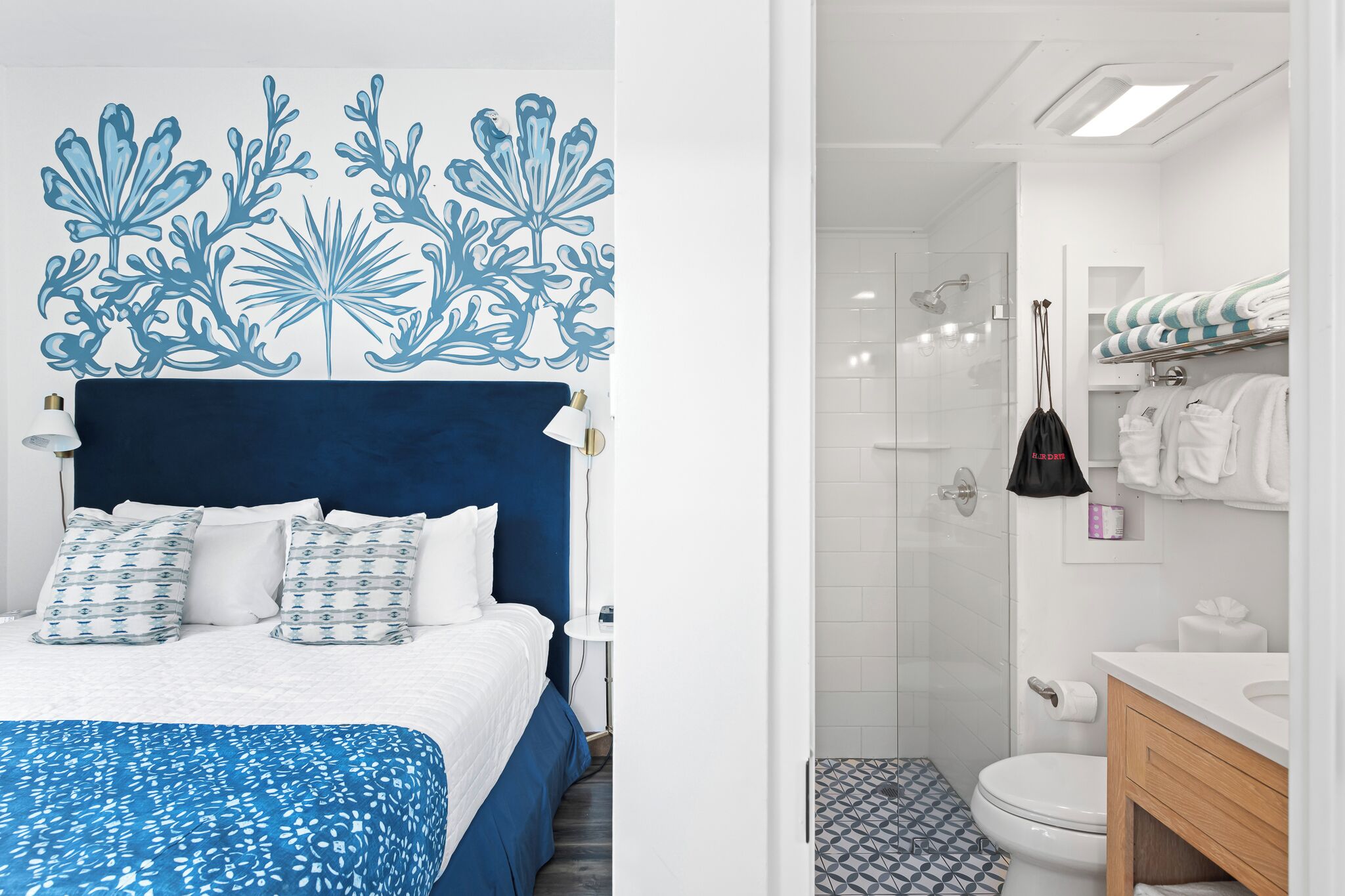 Un dormitorio con cabecera azul, ropa de cama blanca y un adorno mural estampado. Junto a él, un baño con ducha, inodoro y sanitarios blancos.