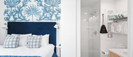 Un dormitorio con cabecera azul, ropa de cama blanca y un adorno mural estampado. Junto a él, un baño con ducha, inodoro y sanitarios blancos.