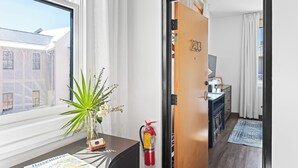 Habitación de hotel con un ventanal grande, una planta en el alféizar y un extintor. Se observa también una puerta con un número.