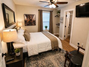 A bedroom with a bed, bedside lamp, a ceiling fan, a TV, a framed picture, a window with curtains, and a door leading to another room.