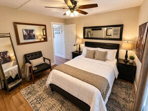 A bedroom with a bed, a chair, a nightstand, a lamp, a ceiling fan, and a door.