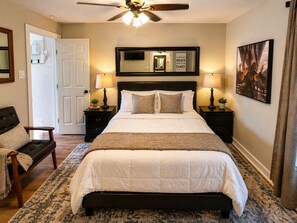 A bedroom with a bed, two bedside tables with lamps, a chair, a ceiling fan, a door, and a framed picture on the wall.