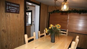 A wooden dining table with a vase of sunflowers, surrounded by wooden chairs. A sign on the wall reads "WILDERNESS RESORT ROUGHING IT IN STYLE." - Triple R Cottages: 1 (Estes Park)