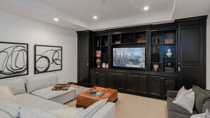A modern living room with a large flat-screen TV, dark wood cabinets, and light grey sofas.