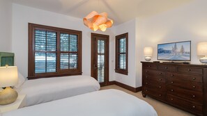 A hotel room with two beds, a large window with blinds, a wooden dresser, a television, and a unique ceiling light fixture.