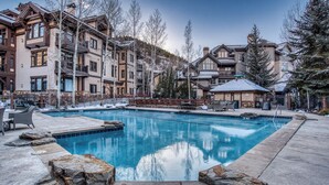 A swimming pool surrounded by multi-story buildings with balconies, a wooden deck, and a snow-covered landscape.