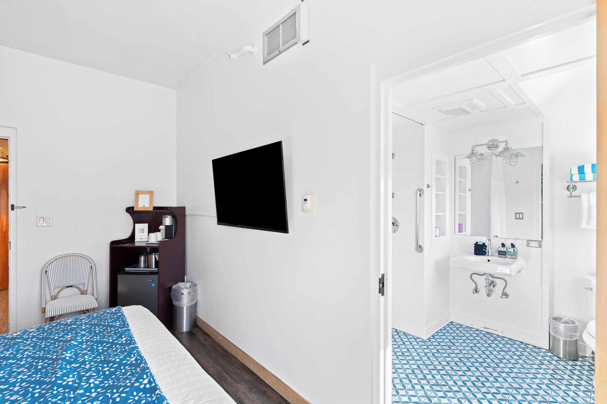 Ett hotellrum med en säng, ett skrivbord med en stol, en TV, ett badrum med handfat och spegel samt ett golv med ett mönster.