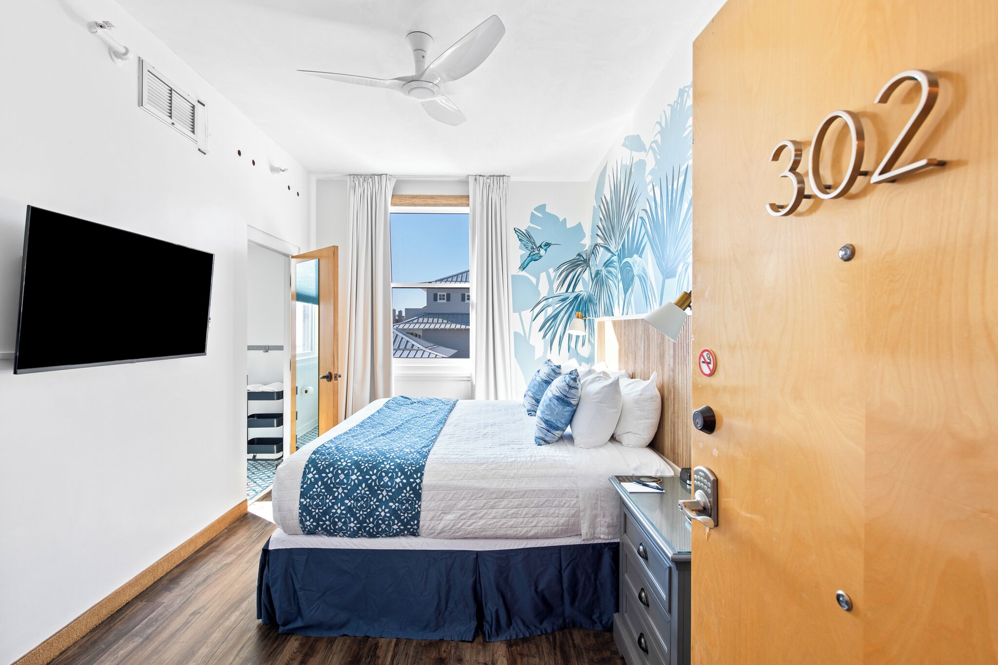 A hotel room with a bed, a TV, a ceiling fan, a window with curtains, and a door with a number.