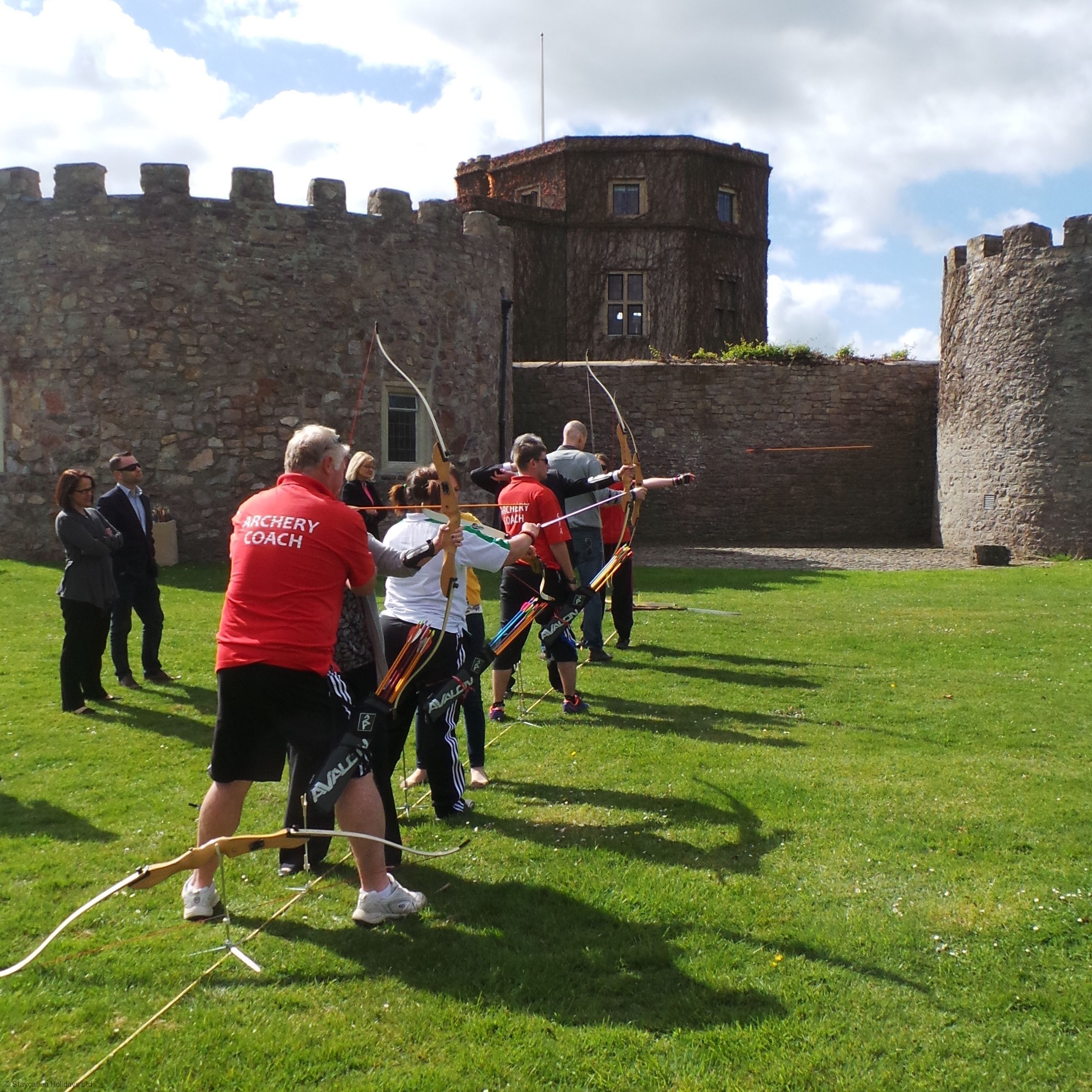 A group of people engaged in archery practice outdoors with a stone building in the background.