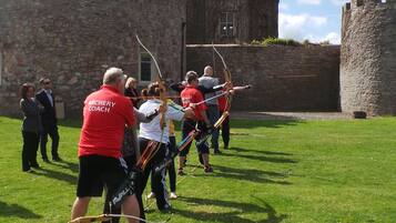 A group of people engaged in archery practice outdoors with a stone building in the background.