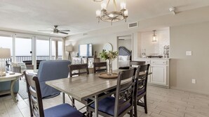 A dining area with a wooden table, blue upholstered chairs, a chandelier, and a view of the balcony. - Orange Beach Gulf-Front 3BR • Sleeps 10 • Bunk Room • Games • Oversized Balcony (Orange Beach)