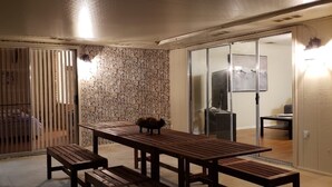 Une pièce avec un mur en pierre, une porte-fenêtre coulissante, une table en bois avec des bancs, et une petite table avec une chaise.