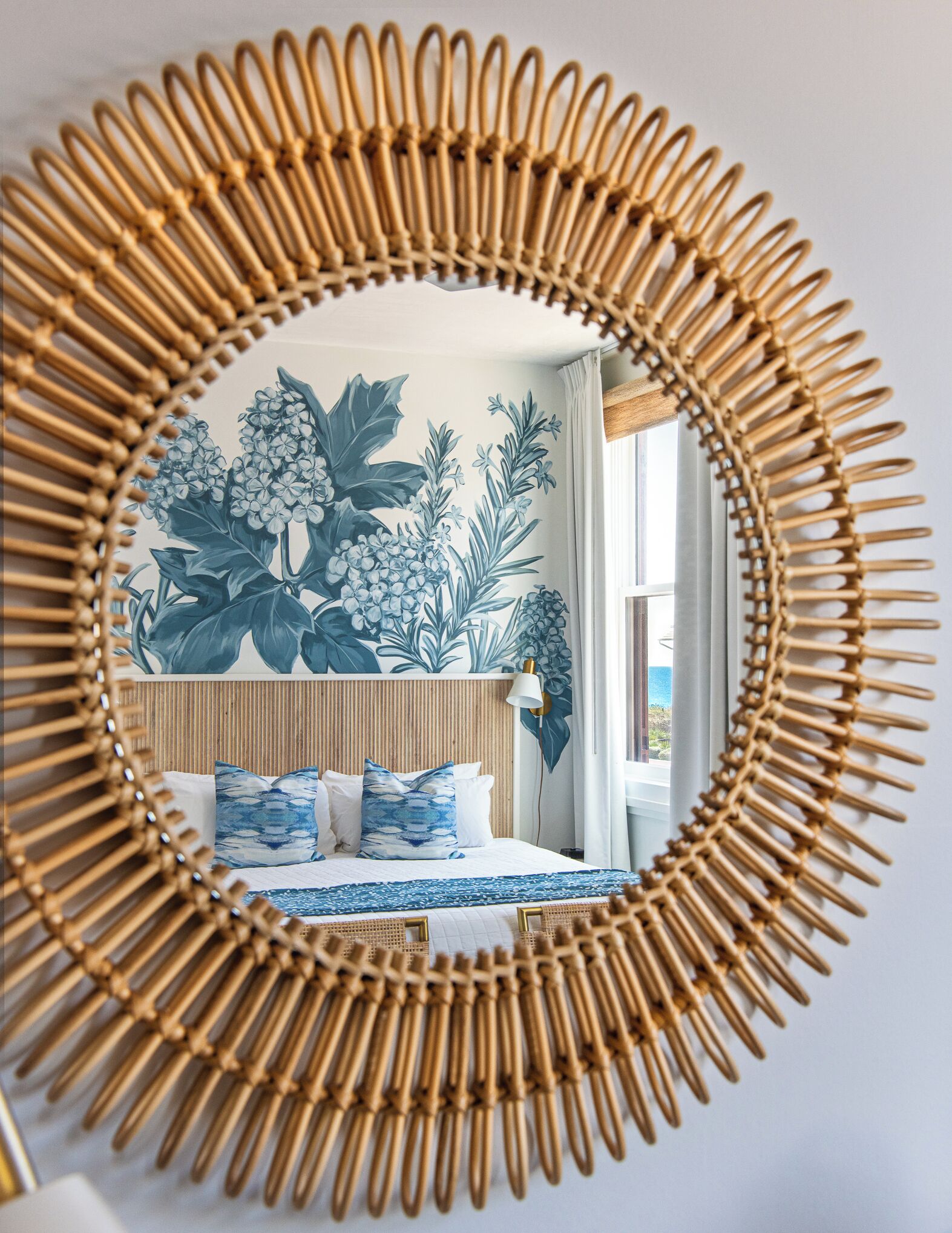 Espelho redondo com moldura de bambu refletindo um quarto com cama, almofadas e um mural na parede.