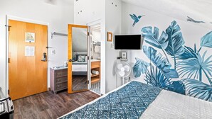 Un dormitorio con puerta de madera, una cama con cubrecama estampado, un televisor de pared, un ventilador y una pared decorada con motivos de hojas tropicales y pájaros.