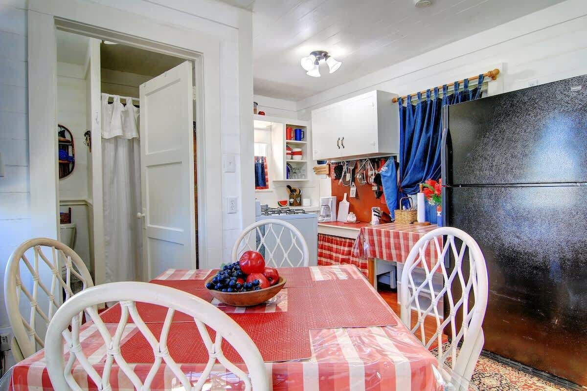 Un comedor con una mesa preparada para dos personas, un cuenco de fruta y una cocina al fondo.