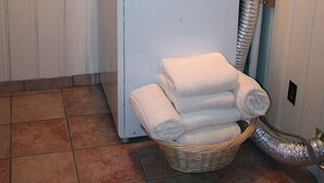 A compact laundry room with a stacked washer and dryer, a basket of folded towels, and a visible water pipe. - Timber Creek Chalets: 14 (Estes Park)
