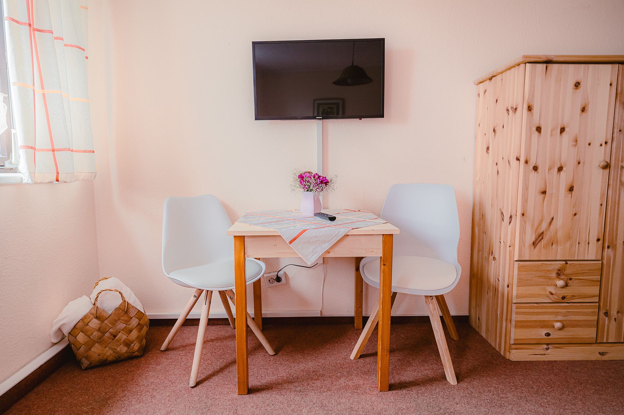 Una habitación con una mesa de madera, dos sillas blancas, un televisor montado en la pared, un armario de madera y un bolso tejido en el suelo.