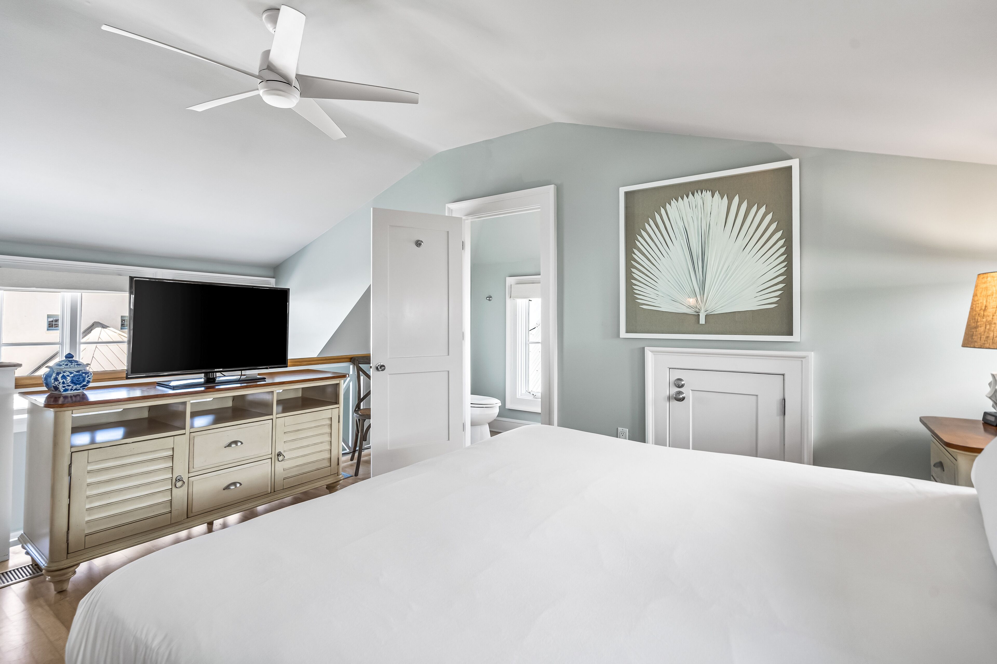 A bedroom with a large bed, a ceiling fan, a TV, a dresser, and a framed artwork on the wall.