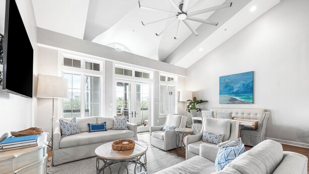 A spacious living room with a high ceiling, large windows, and a modern ceiling fan.