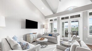 A spacious living room with a grey sofa set, a flat-screen TV mounted on the wall, and a large window with a view of the outdoors.