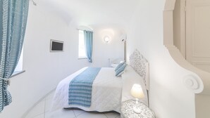 A bedroom with a bed, a nightstand, a lamp, a window with curtains, and a wall-mounted light fixture. - Villa dei lecci Indaco - Luxury villa on the sea with private jacuzzi (Forio)