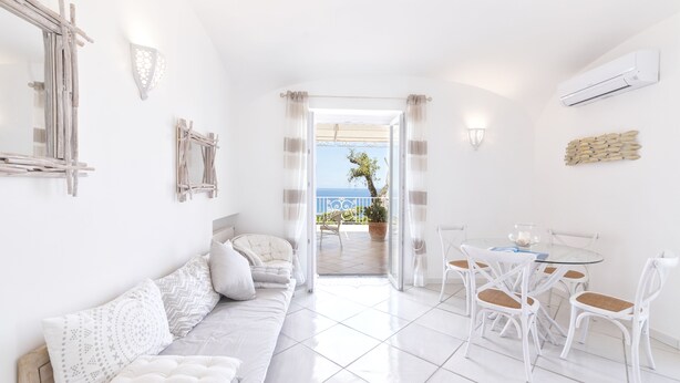 A bright, white room with a sofa, dining table, and chairs. A balcony with a view of the sea is visible through an open door. - Villa dei lecci Indaco - Luxury villa on the sea with private jacuzzi (Forio)