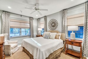 Un dormitorio con una cama grande, ventilador de techo, mesita de noche con lámpara y ventana con cortinas.