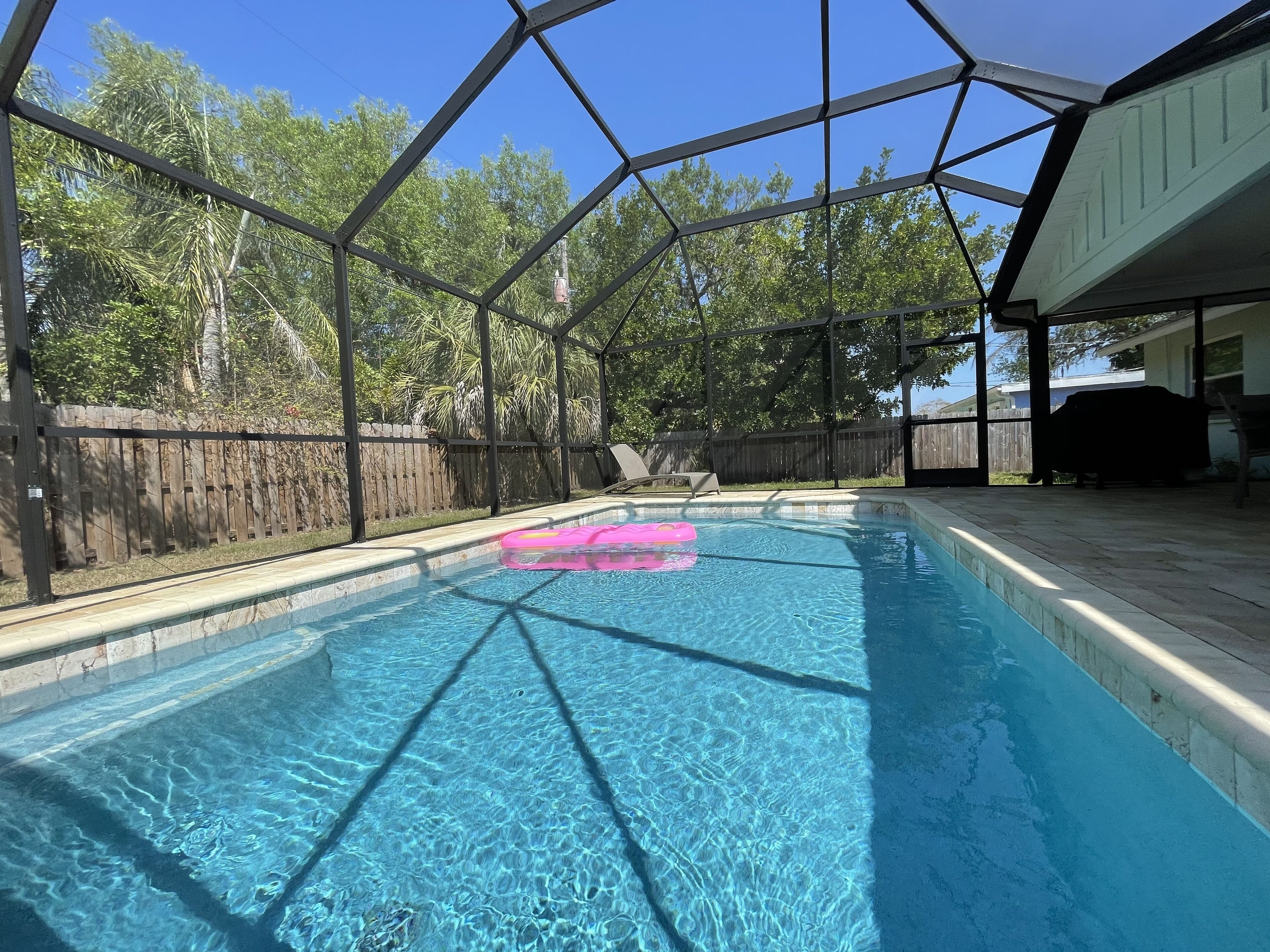A clear blue pool with a pink float, surrounded by a fenced area with a wooden fence and trees in the background.