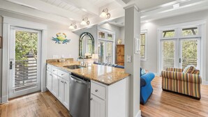 La cuisine est dotée de meubles blancs, d’un plan de travail en granit et d’un lave-vaisselle. Le salon comprend un canapé bleu et un fauteuil à rayures.