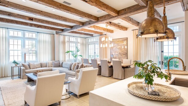 A spacious living area with a large sofa, dining table, and chairs, wooden beams on the ceiling, and large windows.
