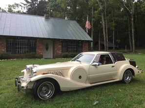 A vintage car parked on grass in front of a building with a brick facade and a green roof.