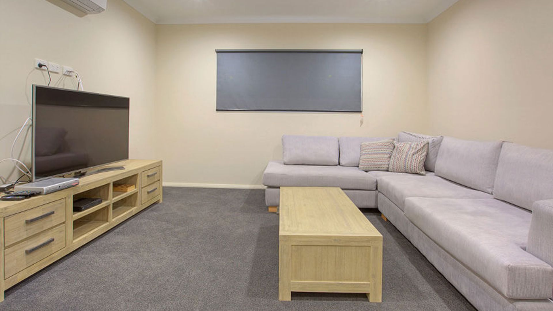 A modern living room with a flat-screen TV, wooden entertainment unit, grey sofa, and a light wooden coffee table.