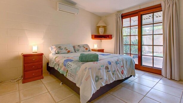 Een slaapkamer met een bed, twee nachtkastjes, een lamp, een raam met gordijnen en een wandairconditioner.