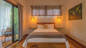 A bedroom with a large bed, wooden headboard, two bedside tables with lamps, a window with curtains, and a painting on the wall. - El Nido Jungle Lodge (Cahuita)