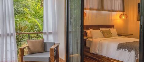Un dormitorio con piso de madera, una cama con ropa de cama blanca, una mesita de noche y un ventanal con cortinas.