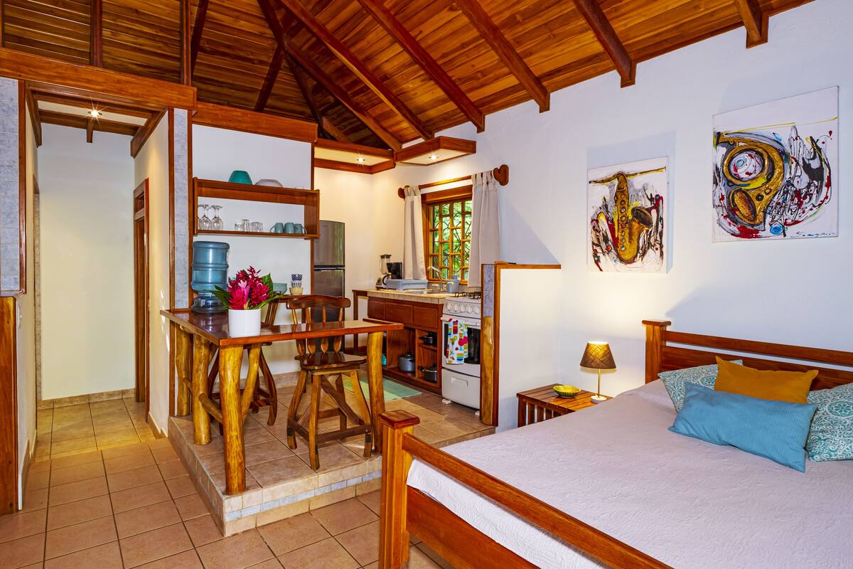 A cozy room with a wooden bed, a dining table with chairs, a kitchen area, and artwork on the walls.