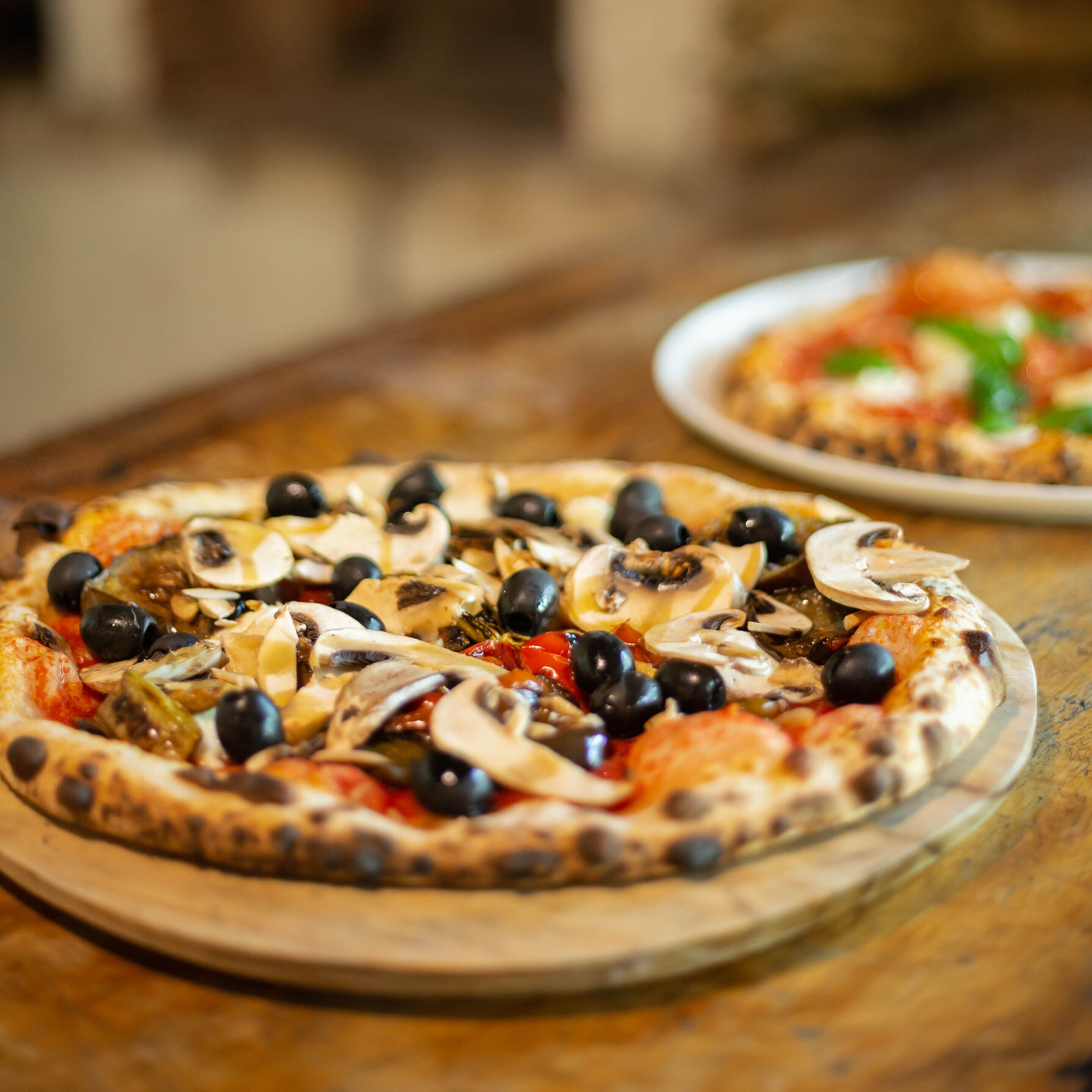 Two pizzas on wooden boards with toppings like olives, mushrooms, and tomatoes.