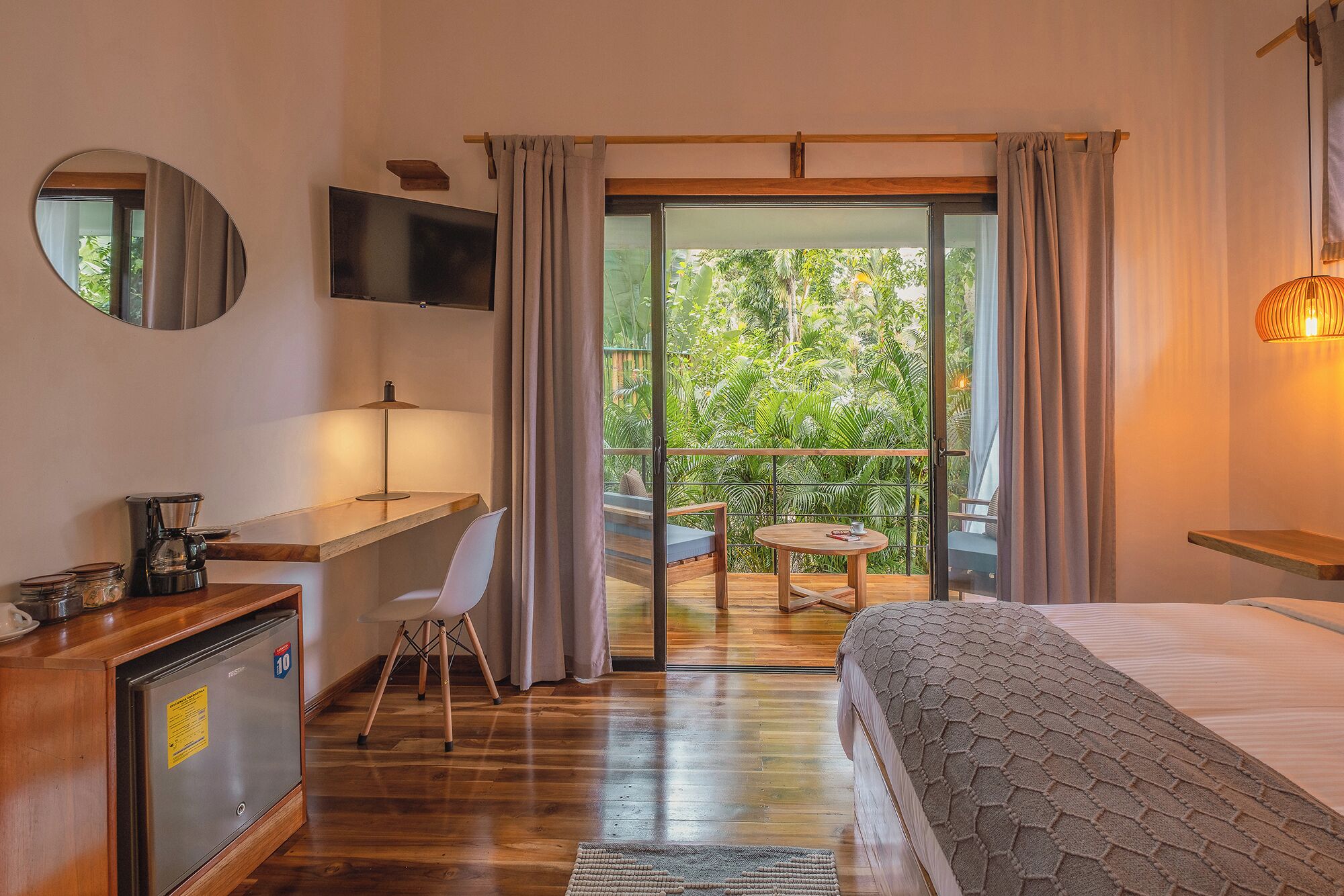 Ein Hotelzimmer mit Bett, Schreibtisch, Sessel, Fernseher, Kaffeemaschine und einem Balkon mit Blick auf Grünflächen.