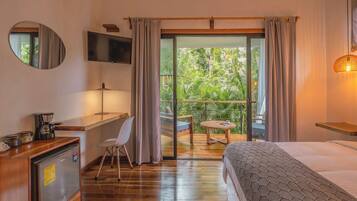 Ein Hotelzimmer mit Bett, Schreibtisch, Sessel, Fernseher, Kaffeemaschine und einem Balkon mit Blick auf Grünflächen.
