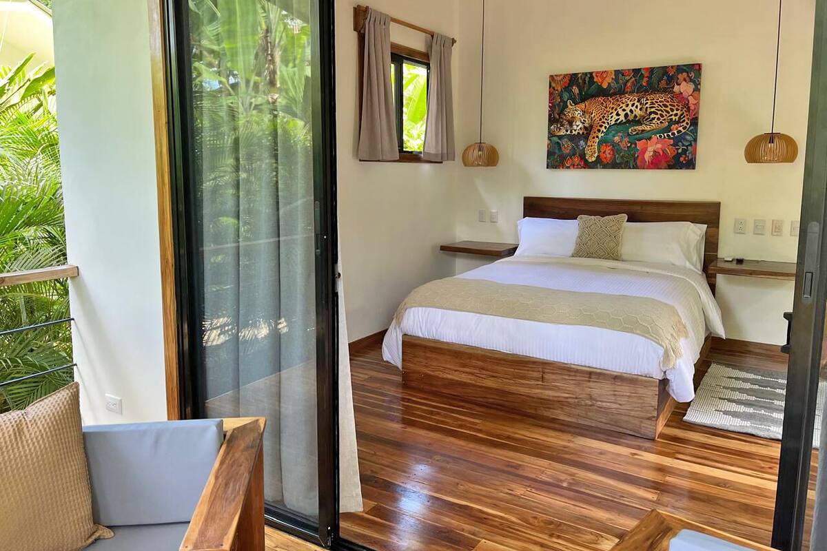 A bedroom with a bed, a wooden floor, a sliding glass door, a painting, and a hanging light fixture.