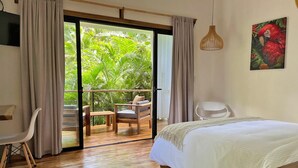 A bedroom with a bed, a chair, a desk, and a balcony with a view of greenery. - El Nido Jungle Lodge (Cahuita)