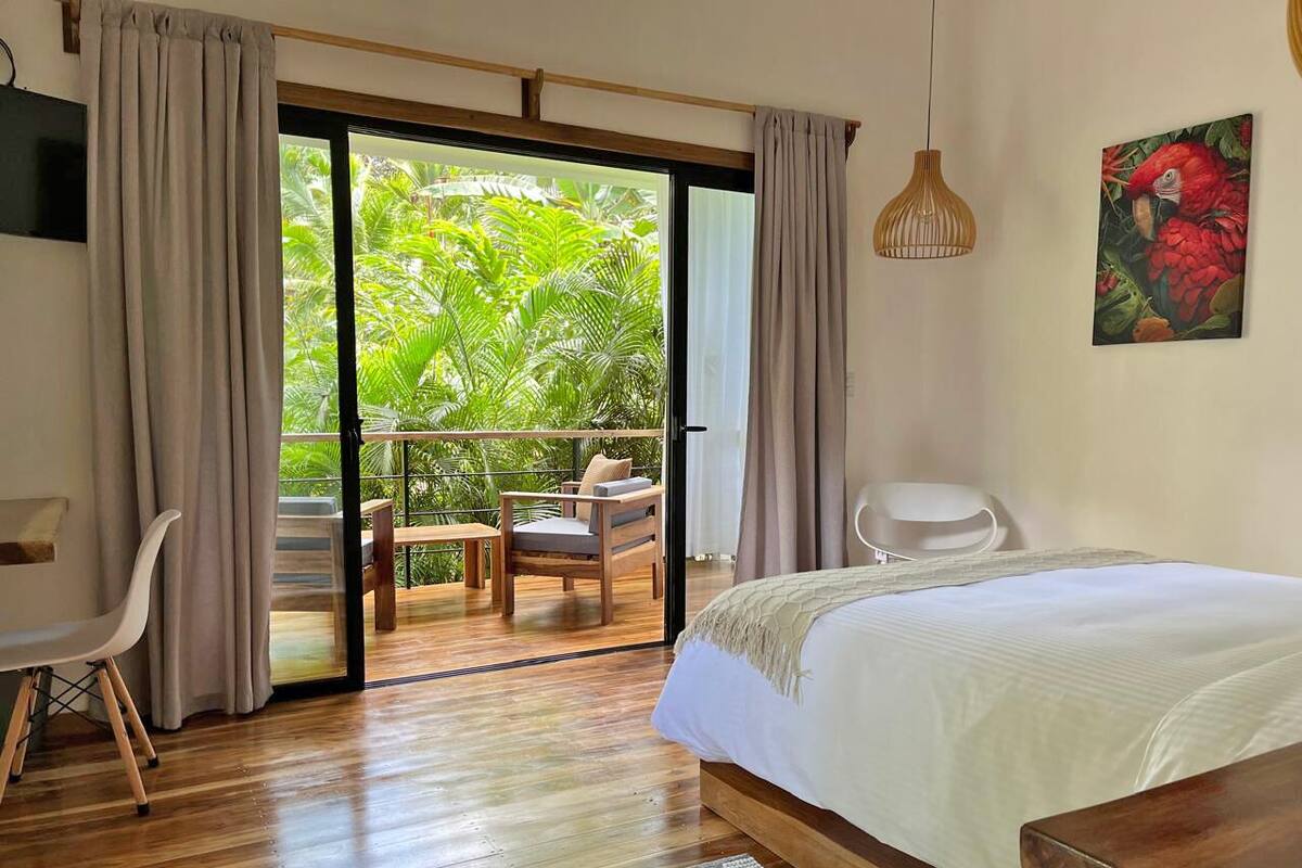 A bedroom with a bed, a chair, a desk, and a balcony with a view of greenery.