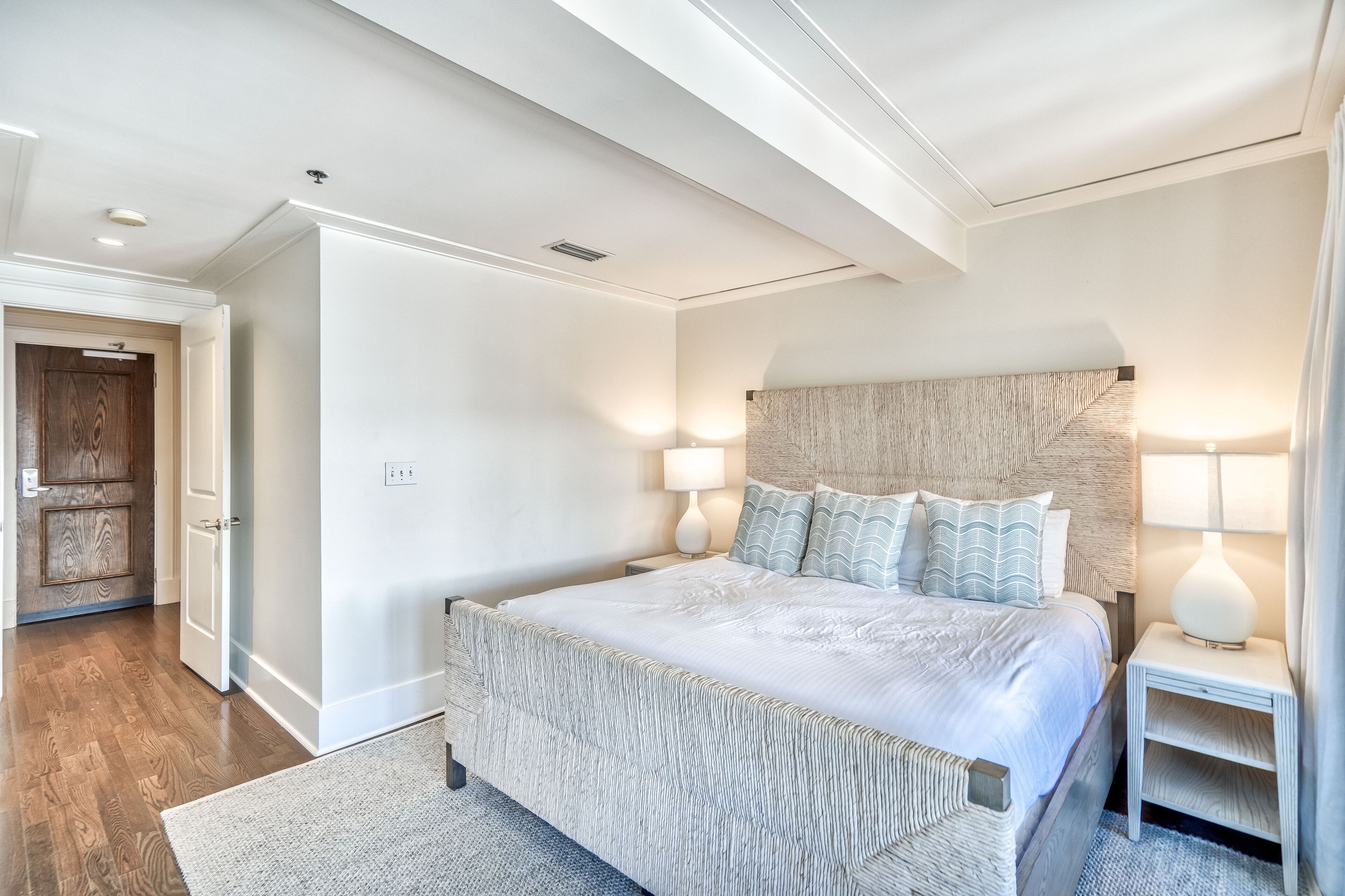 A bedroom with a large bed, two bedside tables with lamps, a wooden door, and a ceiling with recessed lighting.