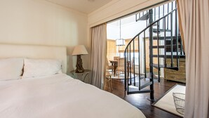 A hotel room with a large bed, a small round table with a lamp, and a view of a spiral staircase leading to a balcony. - Mini Beach Bungalow CH - GULF VIEWS, SOUTH OF 30A. FREE BIKES/GYM/FITNESS (Rosemary Beach)