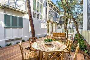 Eine Holzterrasse mit rundem Tisch und Stühlen, umgeben von Grünpflanzen und Bäumen.