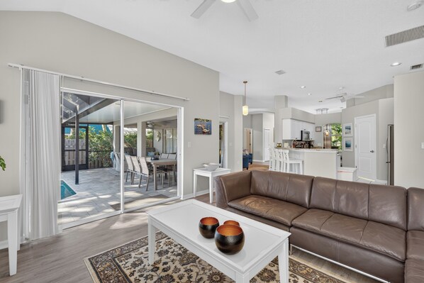 A spacious living area with a brown leather sofa, a white coffee table with decorative bowls, and a dining area with a glass door leading to a patio.
