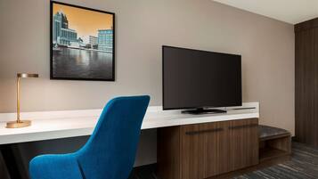 Habitación de hotel con una televisión de pantalla plana sobre una consola blanca, una silla azul, un cuadro enmarco con una vista urbana y una lámpara de pie.