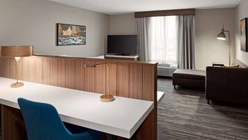 A hotel room with a white desk, a blue chair, a television, a sofa, and a lamp.